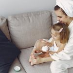 A mother and daughter in bathrobes bonding on a couch, applying moisturizer together.
