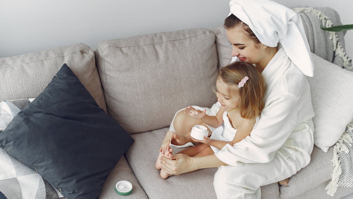 A mother and daughter in bathrobes bonding on a couch, applying moisturizer together.